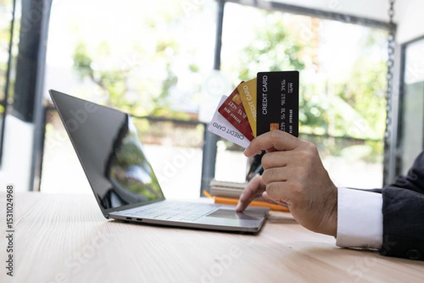 Fototapeta A man is using a laptop and a cell phone to pay for something. He is holding a credit card and a debit card
