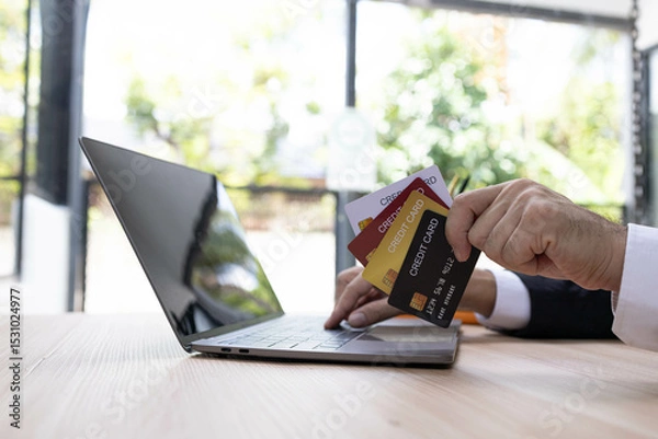 Fototapeta A man is using a laptop and a cell phone to pay for something. He is holding a credit card and a debit card

