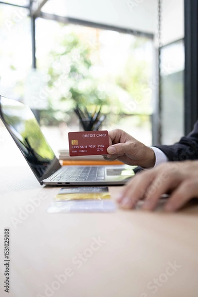 Fototapeta A man is using a laptop and a cell phone to pay for something. He is holding a credit card and a debit card
