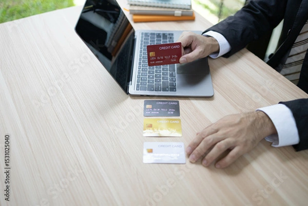 Fototapeta A man is using a laptop and a cell phone to pay for something. He is holding a credit card and a debit card

