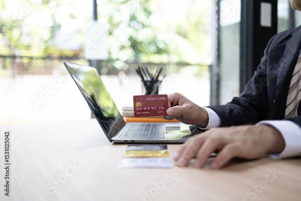 Fototapeta A man is using a laptop and a cell phone to pay for something. He is holding a credit card and a debit card
