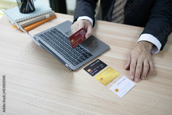Fototapeta A man is using a laptop and a cell phone to pay for something. He is holding a credit card and a debit card
