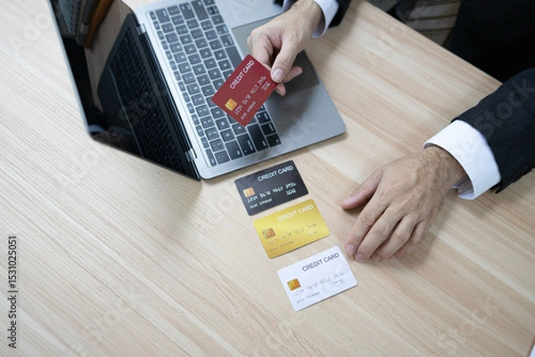 Fototapeta A man is using a laptop and a cell phone to pay for something. He is holding a credit card and a debit card
