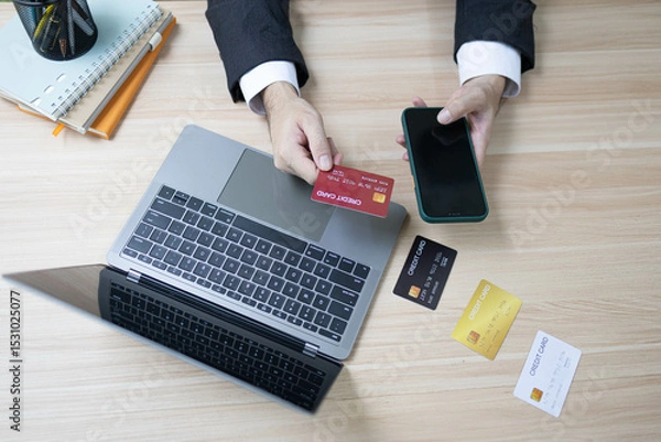 Fototapeta A man is using a laptop and a cell phone to pay for something. He is holding a credit card and a debit card
