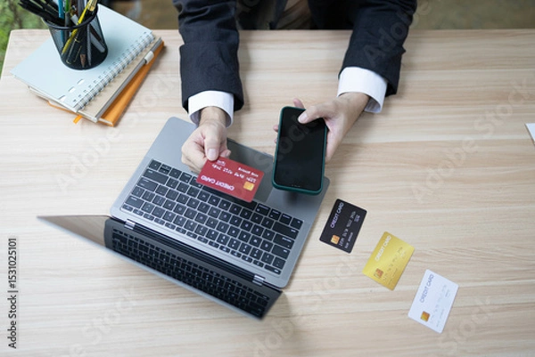 Fototapeta A man is using a laptop and a cell phone to pay for something. He is holding a credit card and a debit card
