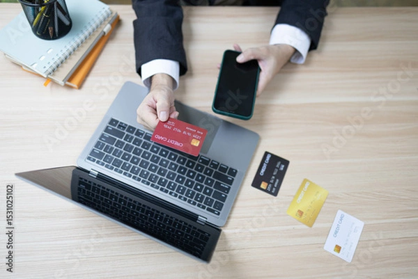 Fototapeta A man is using a laptop and a cell phone to pay for something. He is holding a credit card and a debit card
