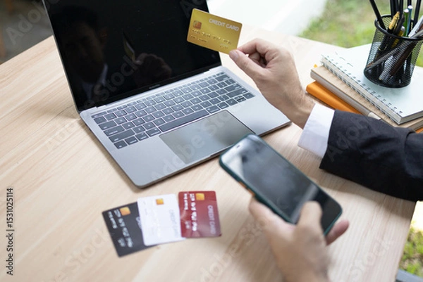 Fototapeta A man is using a laptop and a cell phone to pay for something. He is holding a credit card and a debit card
