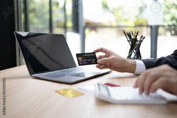 Fototapeta A man is using a laptop and a cell phone to pay for something. He is holding a credit card and a debit card
