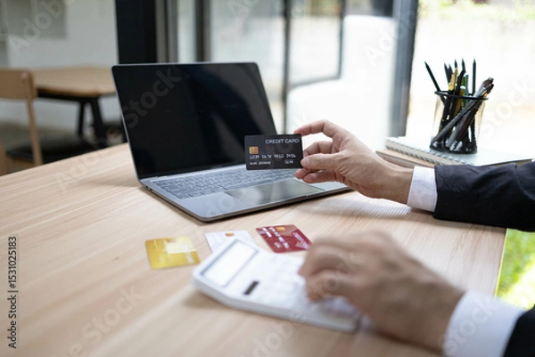 Fototapeta A man is using a laptop and a cell phone to pay for something. He is holding a credit card and a debit card
