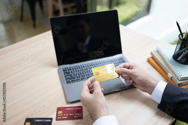 Fototapeta A man is using a laptop and a cell phone to pay for something. He is holding a credit card and a debit card
