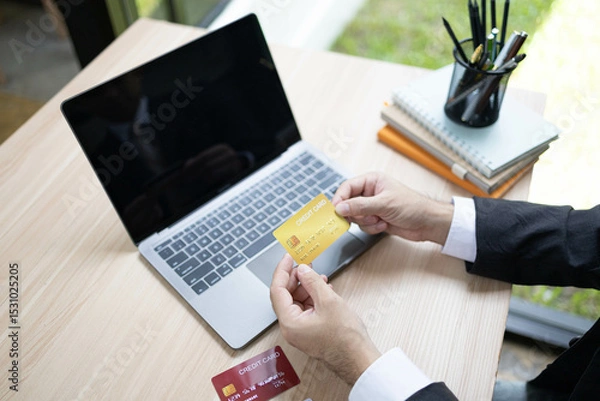 Fototapeta A man is using a laptop and a cell phone to pay for something. He is holding a credit card and a debit card
