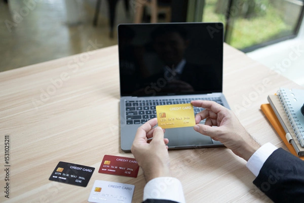 Fototapeta A man is using a laptop and a cell phone to pay for something. He is holding a credit card and a debit card

