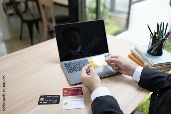 Fototapeta A man is using a laptop and a cell phone to pay for something. He is holding a credit card and a debit card
