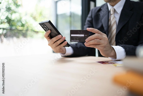 Fototapeta A man is using a laptop and a cell phone to pay for something. He is holding a credit card and a debit card
