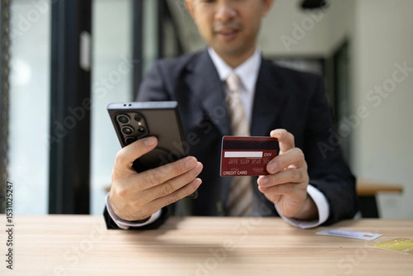 Fototapeta A man is using a laptop and a cell phone to pay for something. He is holding a credit card and a debit card
