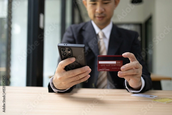 Fototapeta A man is using a laptop and a cell phone to pay for something. He is holding a credit card and a debit card
