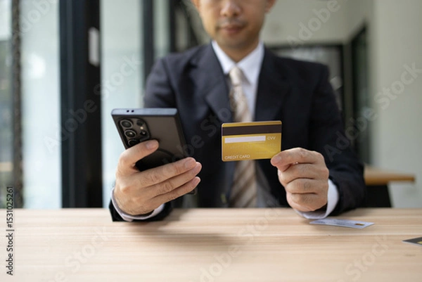 Fototapeta A man is using a laptop and a cell phone to pay for something. He is holding a credit card and a debit card
