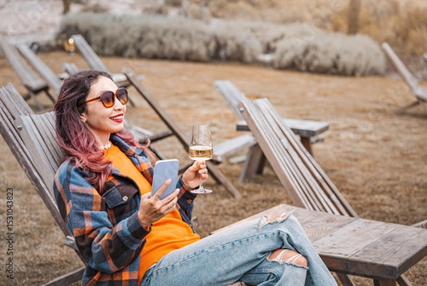 Fototapeta Tourist enjoying a glass of wine while using her smartphone, sitting on a wooden deck chair in a winery garden