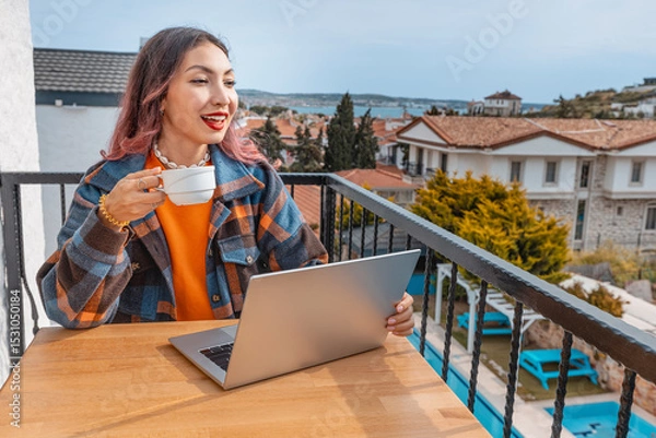 Fototapeta Young woman working remotely on vacation, enjoying a cup of coffee on a balcony with a beautiful sea view