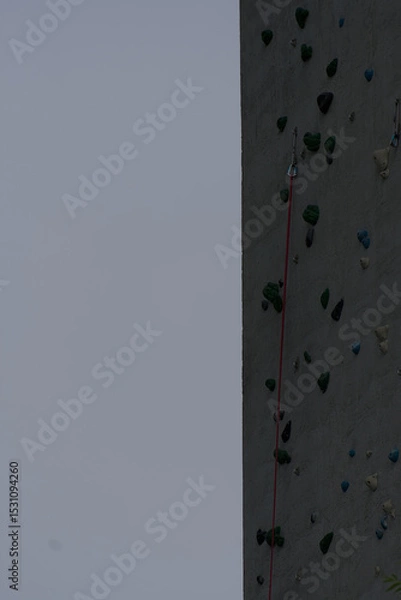 Fototapeta A textured climbing wall with colorful holds and a safety rope extends upward under a muted sky, conveying challenge and vertical adventure