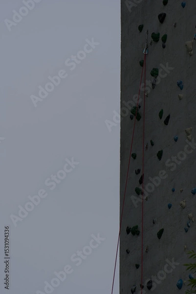 Fototapeta A textured climbing wall with colorful holds and a safety rope extends upward under a muted sky, conveying challenge and vertical adventure