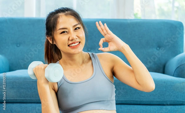 Obraz Young pregnant Asian woman about to become a mother, wearing a sports bra, exercising, and sitting lifting dumbbells up and down slowly to flex her arm muscles before giving birth on a mat, Looks OK