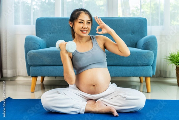 Obraz Young pregnant Asian woman about to become a mother, wearing a sports bra, exercising, and sitting lifting dumbbells up and down slowly to flex her arm muscles before giving birth on a mat, Looks OK