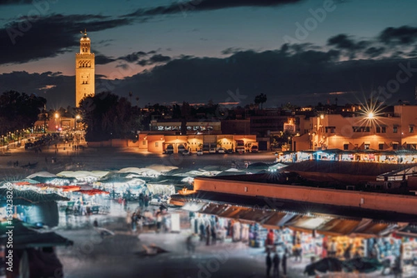 Fototapeta Illuminated stalls and bustling crowds fill Djemaa el Fna market in Marrakech, Morocco, as dusk settles, with the Koutoubia Mosque minaret