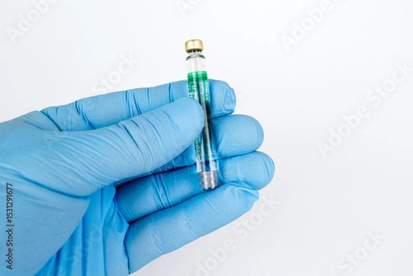 Obraz Gloved Hand Holding Medical Vaccine Vial on White Background, Dental Anesthesia Syringe.Close-up of a blue-gloved hand holding a small glass vaccine vial with a green label