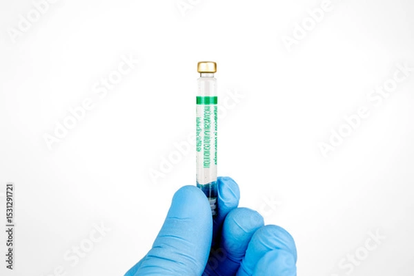 Obraz Gloved Hand Holding Medical Vaccine Vial on White Background, Dental Anesthesia Syringe.Close-up of a blue-gloved hand holding a small glass vaccine vial with a green label