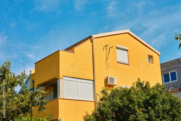 Obraz Yellow house with balconies, roller shutters, and an air conditioner against blue sky. Modern suburban building with bright facade