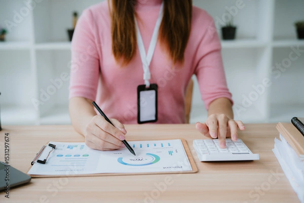 Obraz Young businesswoman calculating and analyzing financial data using calculator and computer while working on charts and graphs in modern office