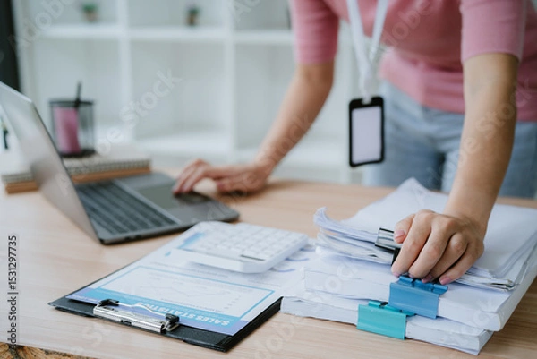 Obraz Woman holding document file stack of business document paper looking at laptop computer flicking through clients files on table, searching for and picking a file in home office.