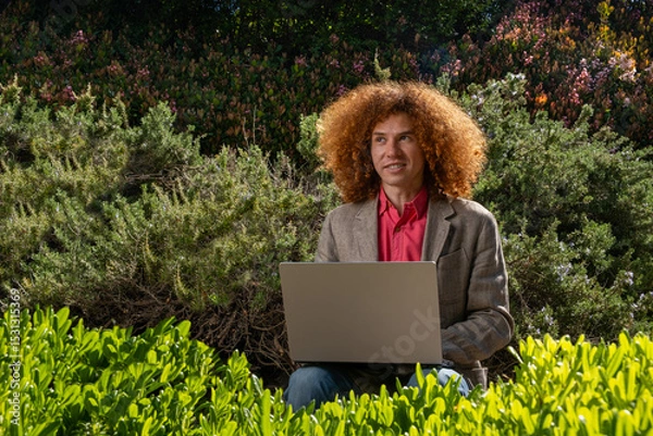 Fototapeta Young man with curly hair working on a laptop outdoors, surrounded by lush greenery. Concept of sustainable remote work, eco-friendly business, and digital nomad lifestyle