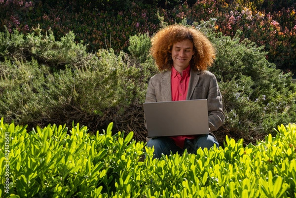 Fototapeta A young executive dressed casually with red afro hair smiles while working. He is working on his laptop in the middle of a public park surrounded by plants. Sustainability and technology