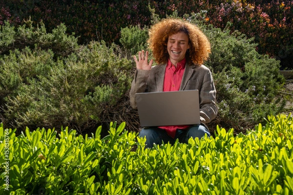 Fototapeta A young redhead executive dressed casually with afro hair waving on a video meeting. He is working on his laptop in the middle of a public park surrounded by plants. Sustainability and technology
