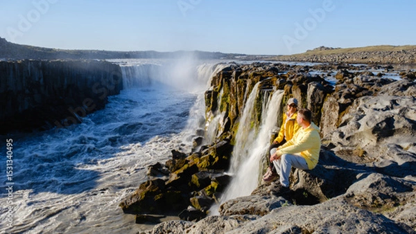 Fototapeta Visitors sit on rocky terrain, captivated by the mighty Dettifoss waterfall in Iceland.