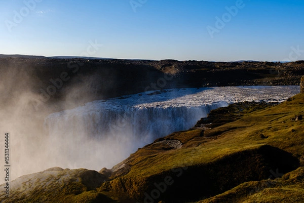 Fototapeta Water plunges powerfully over rocky edges at Dettifss Iceland, creating a misty atmosphere amidst lush greenery.
