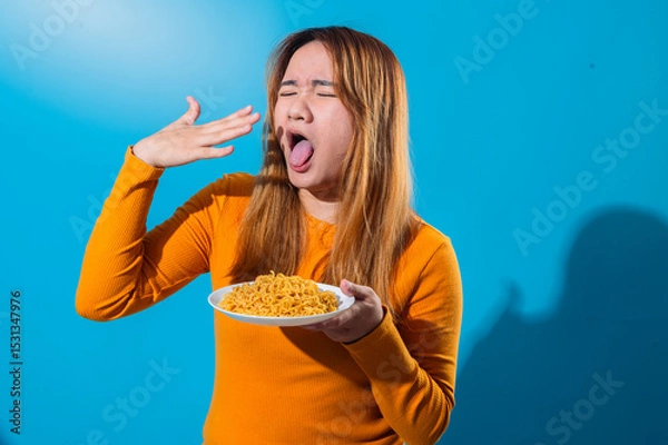 Fototapeta Asian woman in orange shirt with a disgusted expression, sticking out tongue while holding a plate of instant noodles, reacting negatively to the food against a blue background