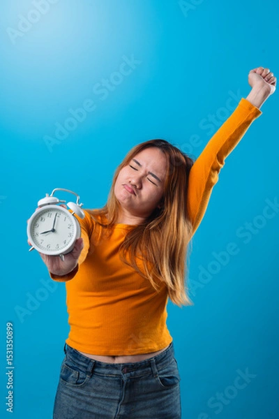 Fototapeta Asian woman stretching with one arm up and holding a white alarm clock forward in the other hand, showing a sleepy expression in front of a solid blue background