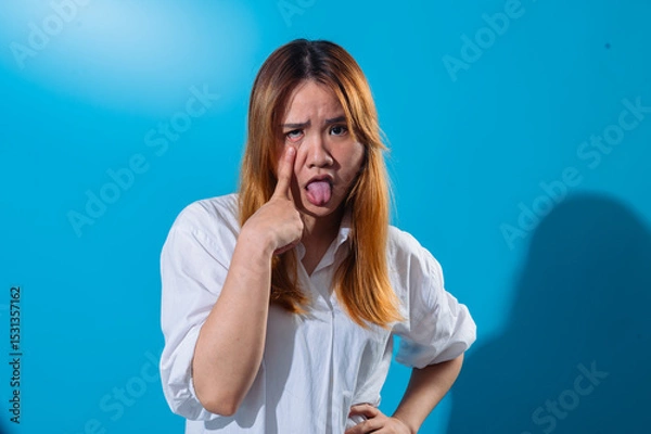 Fototapeta Asian woman in a white shirt pulling her lower eyelid down with one finger and sticking out her tongue in a silly and exaggerated expression, standing against a solid blue background