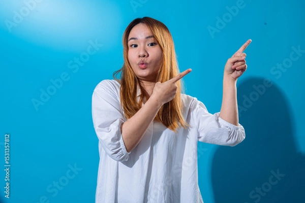 Fototapeta Asian woman with long straight hair wearing casual white shirt smiles brightly while pointing sideways with both hands against vibrant blue background, creating a cheerful and dynamic pose