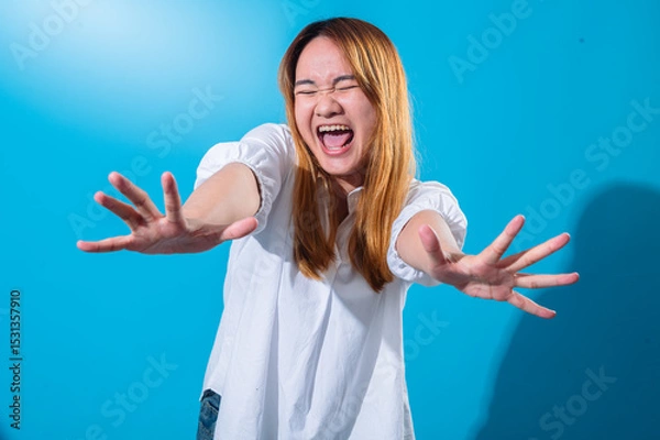 Fototapeta Asian woman with long straight hair in white shirt smiles widely and extends both hands forward in energetic open-palm gesture, standing against solid blue background in expressive studio portrait