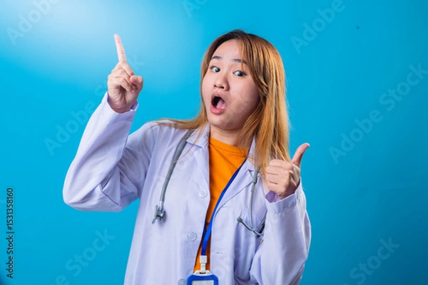 Fototapeta Asian female doctor smiling cheerfully while pointing up with one hand and giving a thumbs up with the other, wearing a white coat and stethoscope on a bright blue background