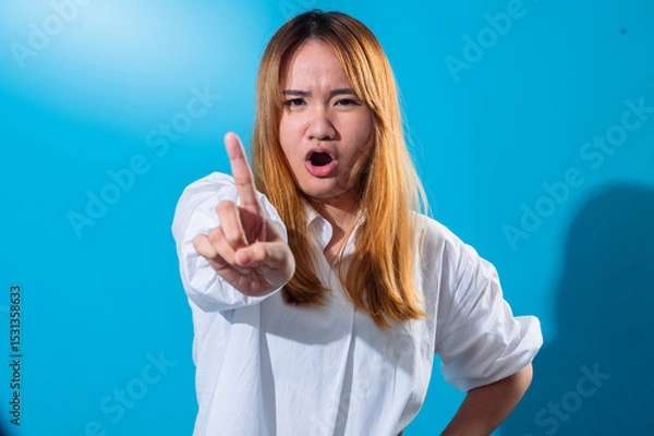 Fototapeta Asian woman in a white shirt pointing her index finger forward with a stern expression, as if warning or scolding, against a plain bright blue background