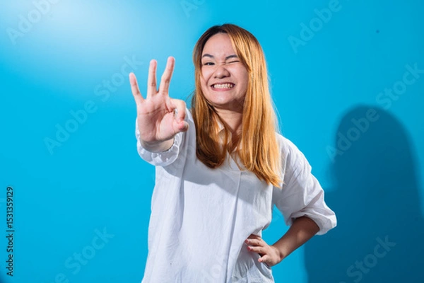 Fototapeta Asian woman with long straight hair in a white shirt smiles brightly while making an OK hand gesture with one hand and winking playfully against a plain blue background in studio lighting