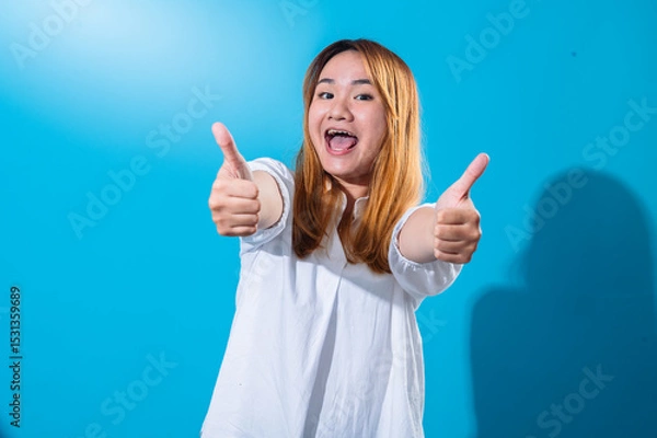 Fototapeta Asian woman with long straight hair in white shirt smiles and enthusiastically gives double thumbs up with extended arms, posing against a solid bright blue background in a cheerful studio setting