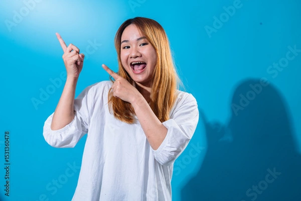 Fototapeta Asian woman with long straight hair wearing casual white shirt smiles brightly while pointing sideways with both hands against vibrant blue background, creating a cheerful and dynamic pose