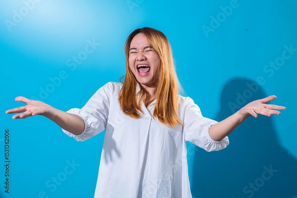 Fototapeta Asian woman with long straight hair wearing white shirt smiles widely while holding both hands open to the sides in welcoming gesture, standing against a bright blue background in studio light