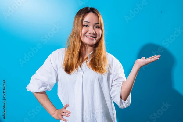 Fototapeta Asian woman with long straight hair in white shirt smiling while holding one hand open to the side as if presenting or showing something, standing confidently against a solid blue studio background